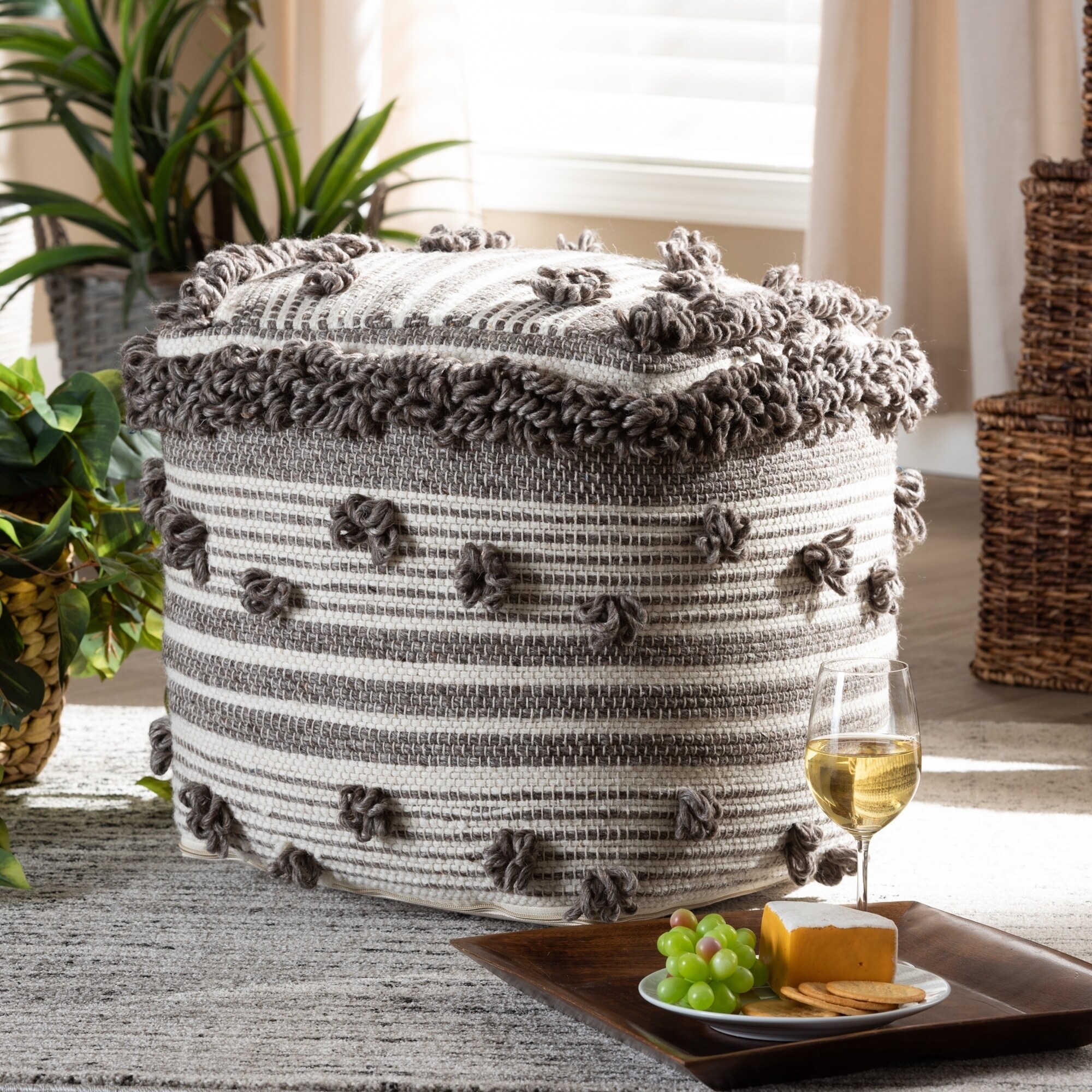 Round woven pouf ottoman with gray and white stripes and fluffy tassel pom pom trim, placed on a rug with plants and wine setup nearby.