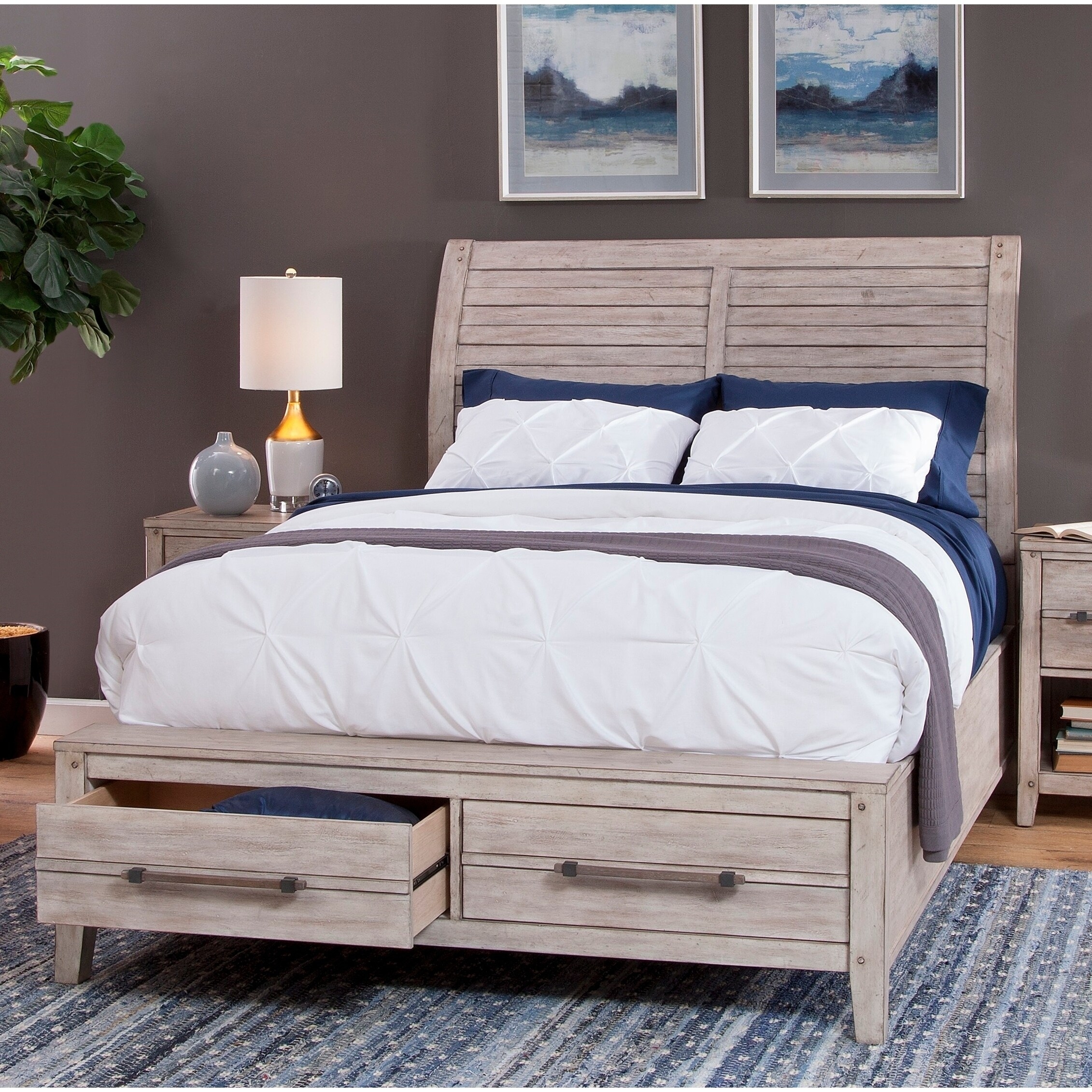 A rustic gray wood bed with built-in under-bed drawers, matching nightstand, dresser, and chest in a bedroom set. White quilted bedding with navy blue accents and a gray throw; lamps and decor on matching tables.