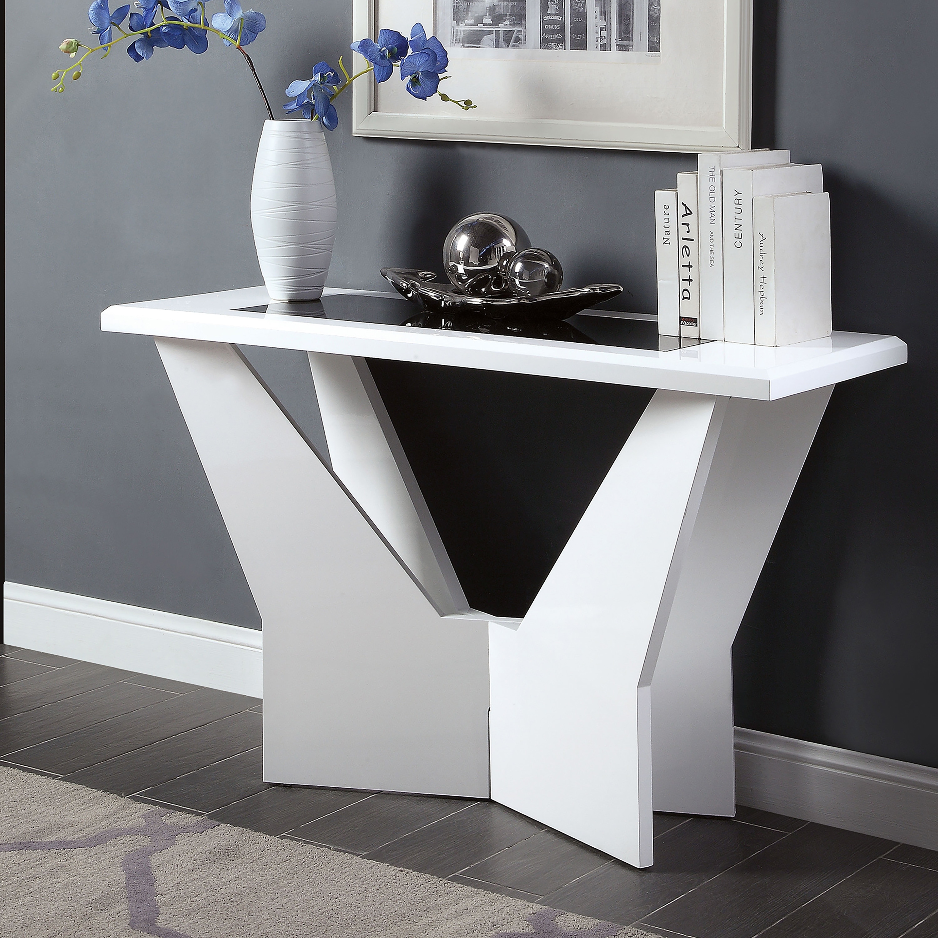 A modern white console table with a geometric base, placed against a dark wall, with a vase of blue flowers, a decorative dish, and a stack of books on top.