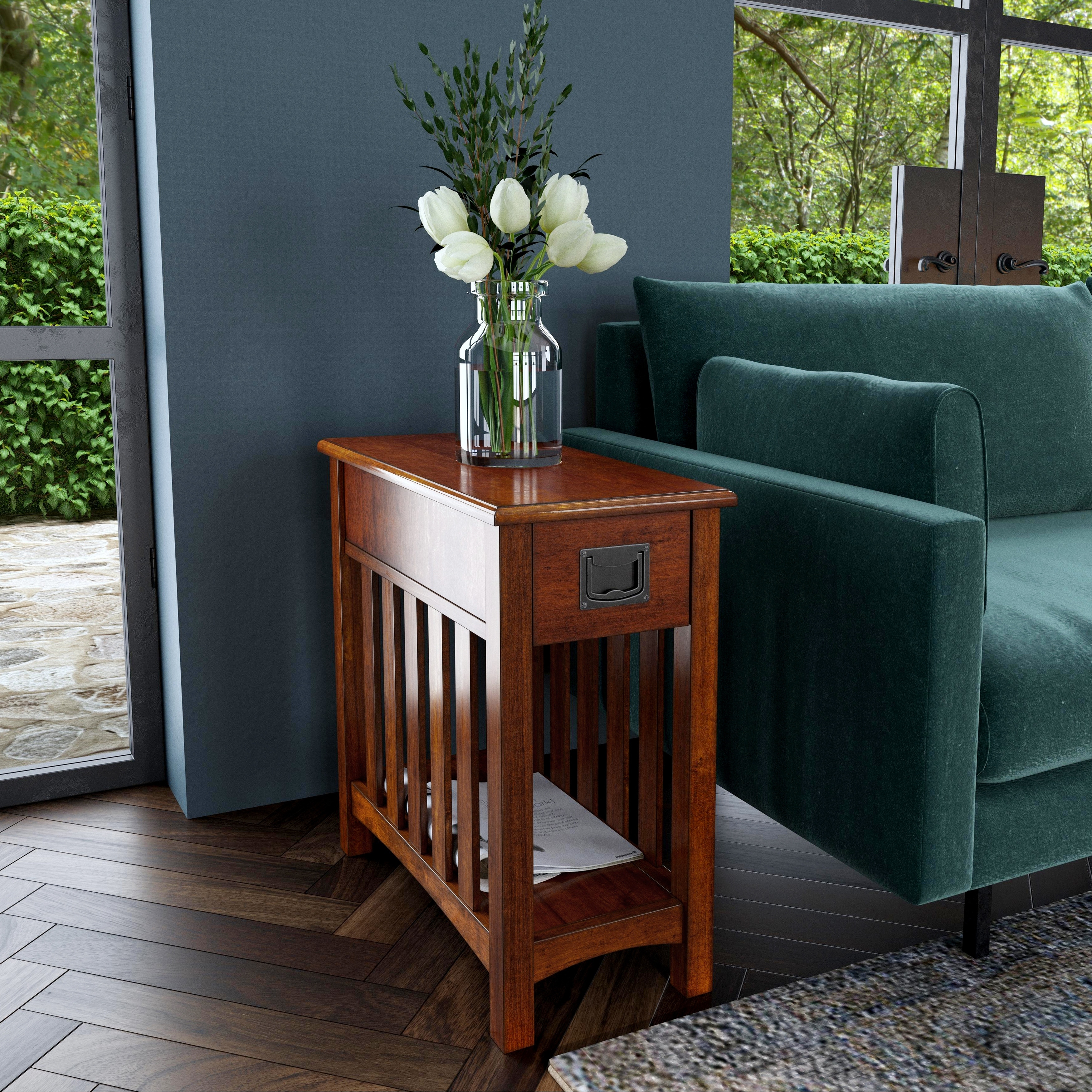 A wooden side table with a drawer and slatted sides placed next to a green sofa; a vase with white flowers sits on top.