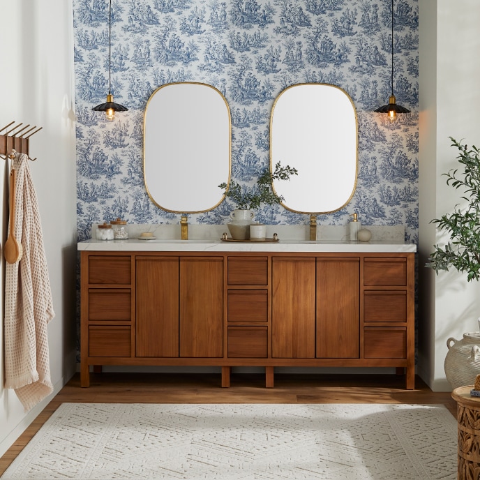 Bathroom with a wooden, marble-topped double vanity against a wall with blue, vintage wall paper and two oval mirrors.