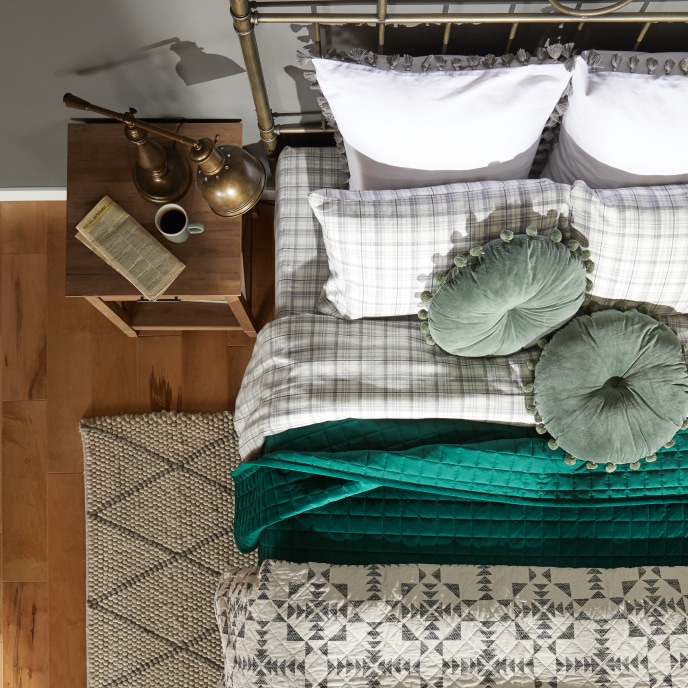 Overhead view of a bed with white and grey bedding, green throw pillows and a coordinating accent blanket, next to a side table with a brass reading lamp.