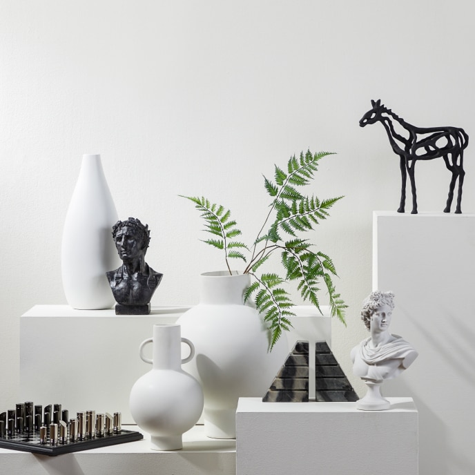 A tiered display of white vases and black sculptures including an abstract horse, a Grecian bust, and a modern chess set.