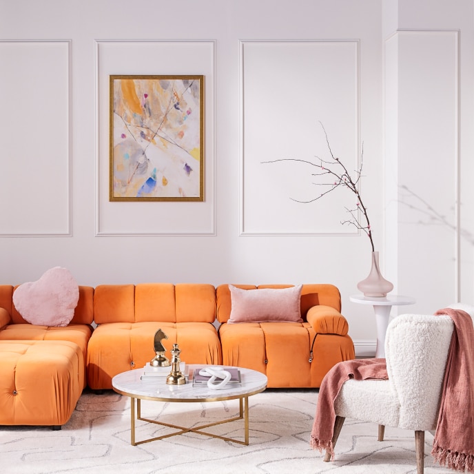 Living room with a modern, orange sectional, chess piece statuettes on a round, gold and marble coffee table, and a white sherpa arm chair with a pink throw blanket.