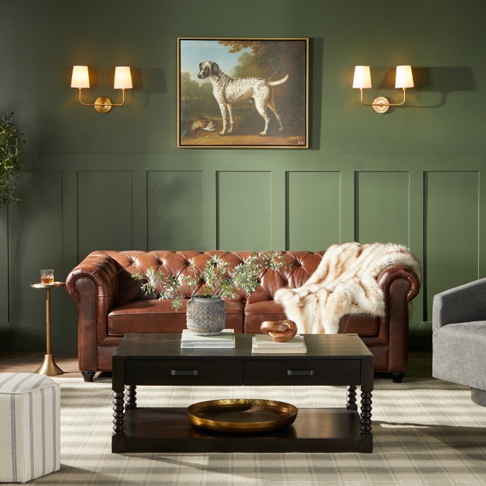 A green living room with a leather sofa with a cozy cream throw blanket and grey, mid-century modern chairs, next to a coffee table with a plant and books.