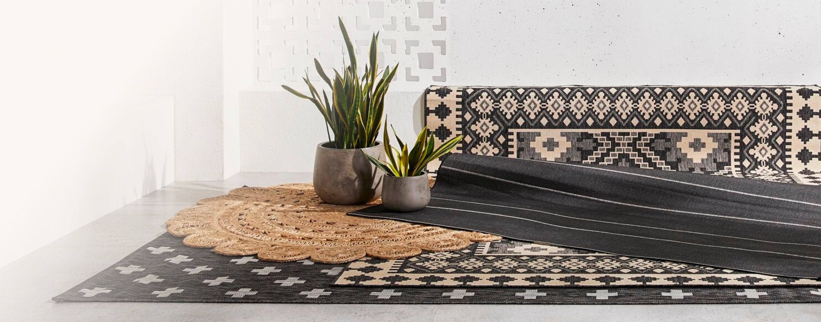 A black rug with a tan persian pattern is laid over a black-and-grey rug. Two potted plants are placed on top of the outdoor rugs.