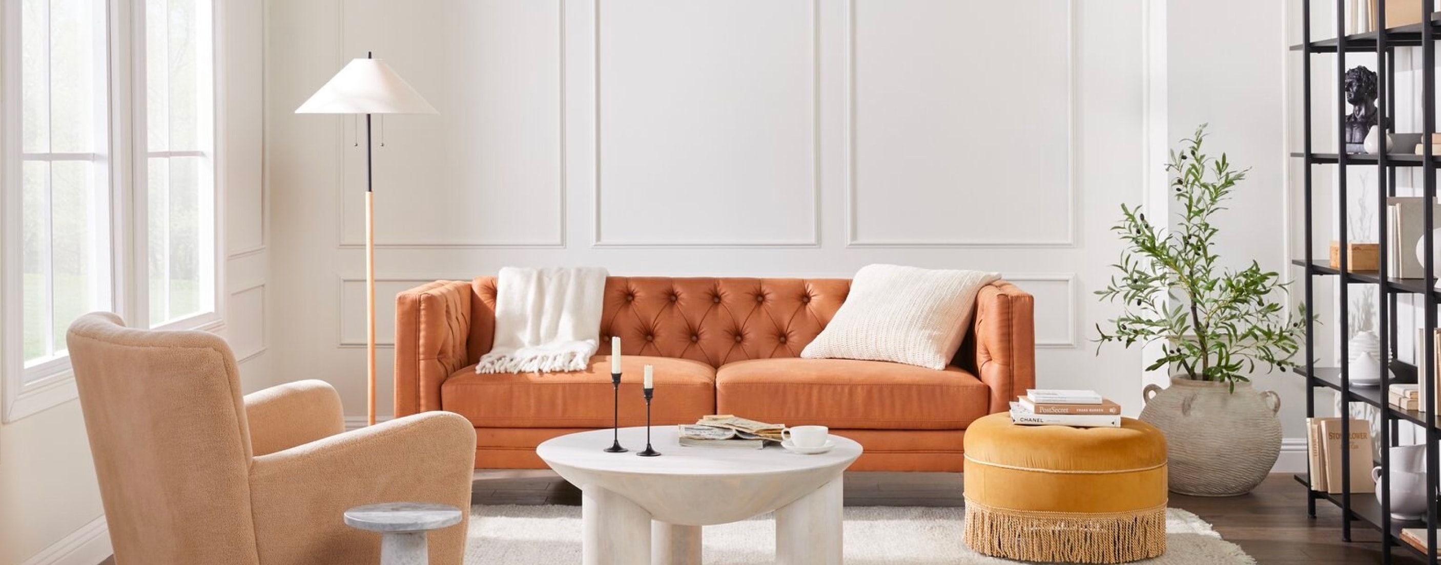 A charming contemporary living room is shown with a burnt orange chesterfield sofa, a peach velvet armchair, a round ottoman, and a marble coffee table.
