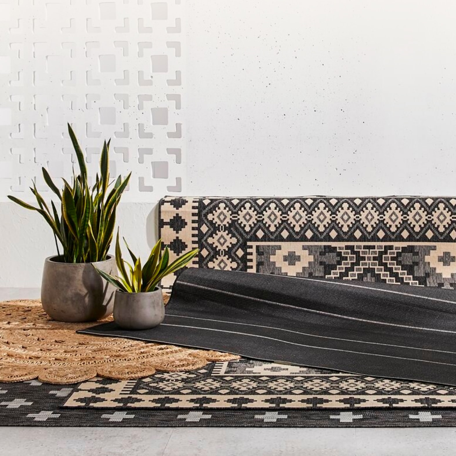 A black rug with a tan persian pattern is laid over a black-and-grey rug. Two potted plants are placed on top of the outdoor rugs.