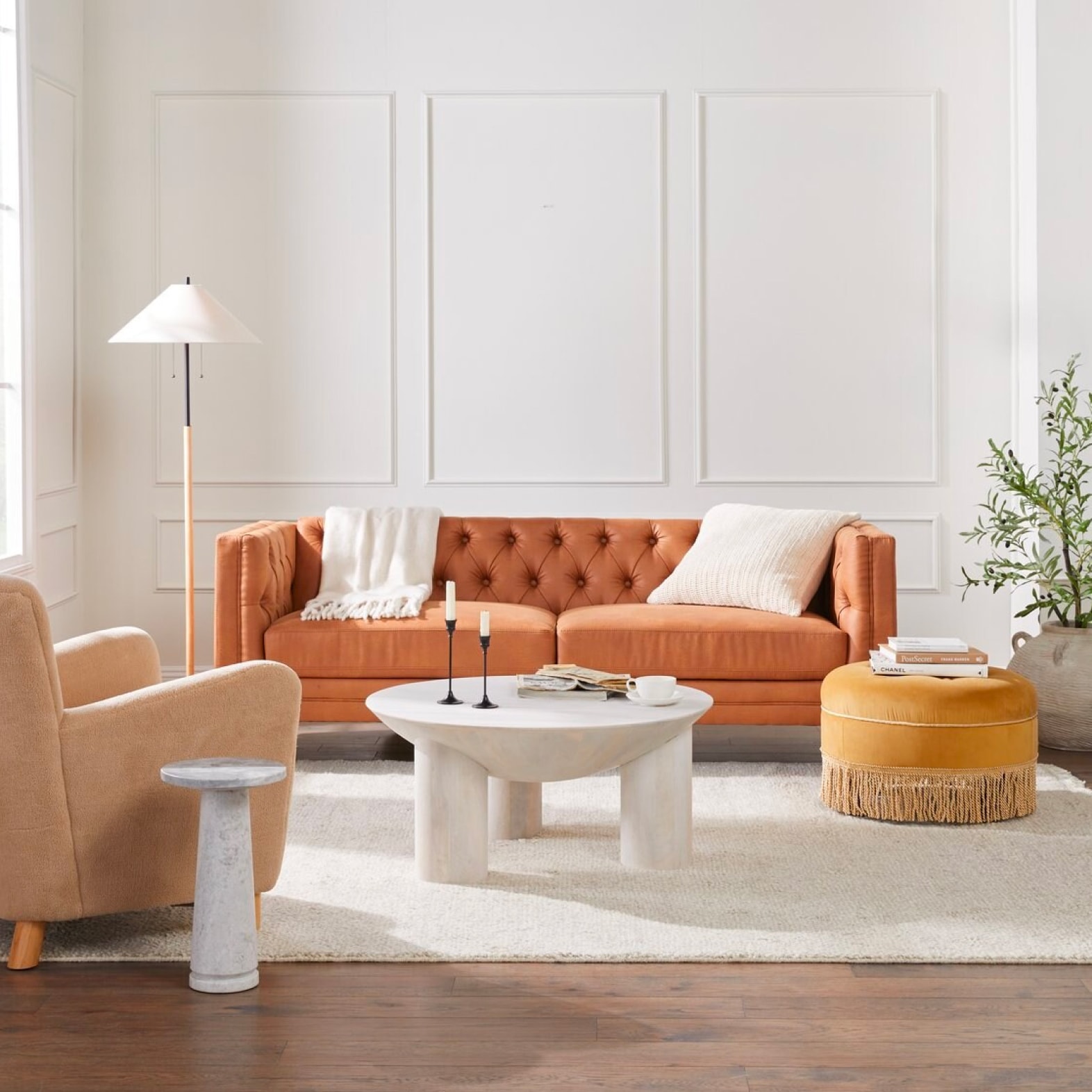 A charming contemporary living room is shown with a burnt orange chesterfield sofa, a peach velvet armchair, a round ottoman, and a marble coffee table.