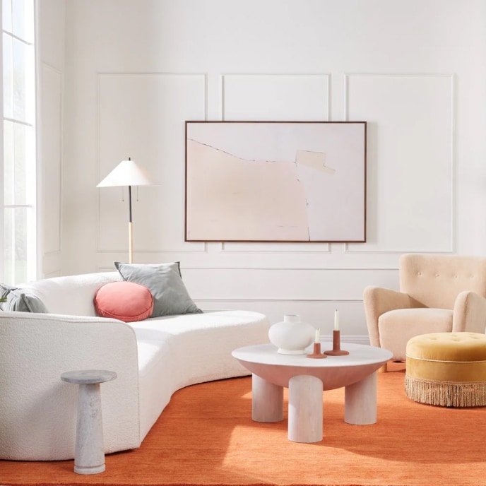 A living room with an orange rug, a curved white sofa, a beige armchair, a yellow ottoman with fringe, and a white statement coffee table topped with candles and a vase.