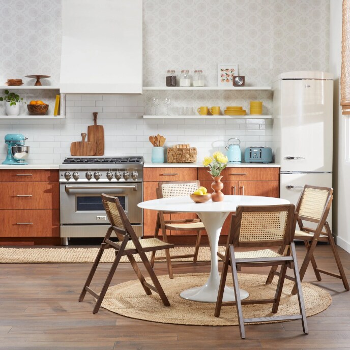 A kitchen with a white tulip table and brown wood and rattan chairs in front of wood cupboards, a range stove, and a retro refrigerator. 