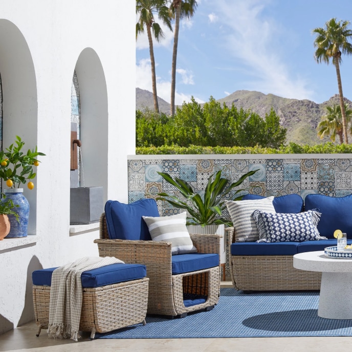 A patio with a wicker seating set with dark blue cushions, near a round white coffee table over a blue area rug.