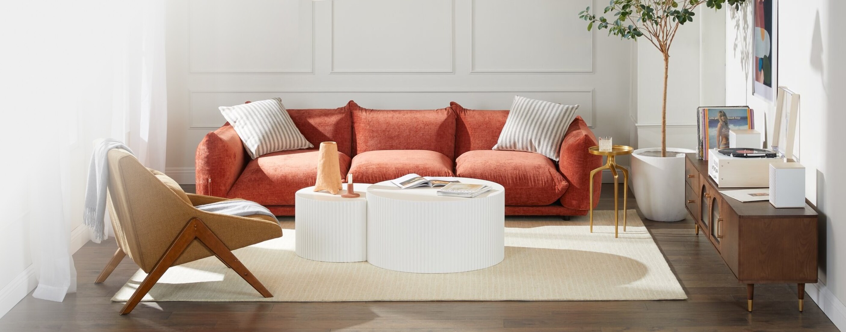 A trendy living room is displayed, featuring a plush sofa with velvety red-orange upholstery, a modern side chair with wood legs, and a two-tiered round white coffee table.