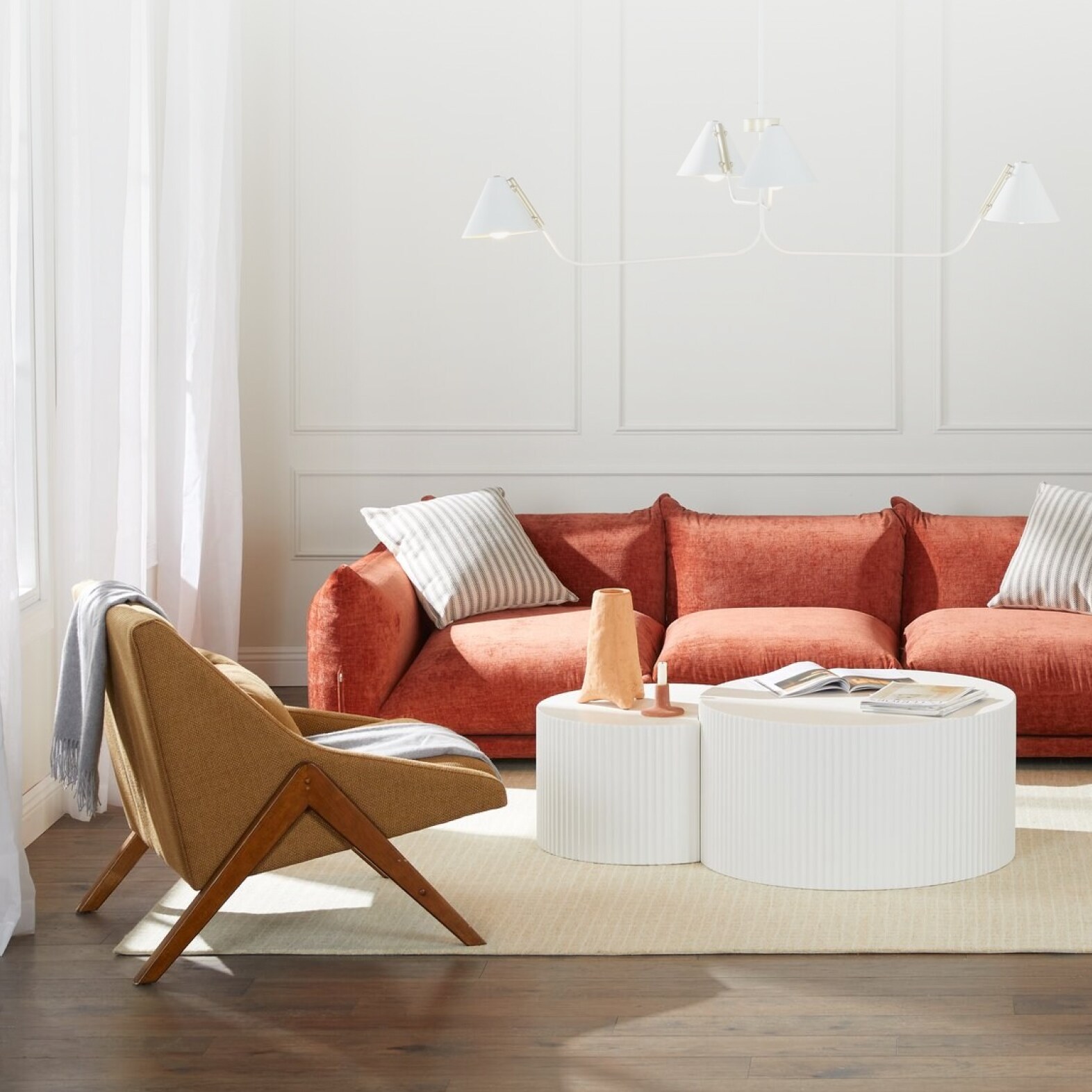 A trendy living room is displayed, featuring a plush sofa with velvety red-orange upholstery, a modern side chair with wood legs, and a two-tiered round white coffee table.