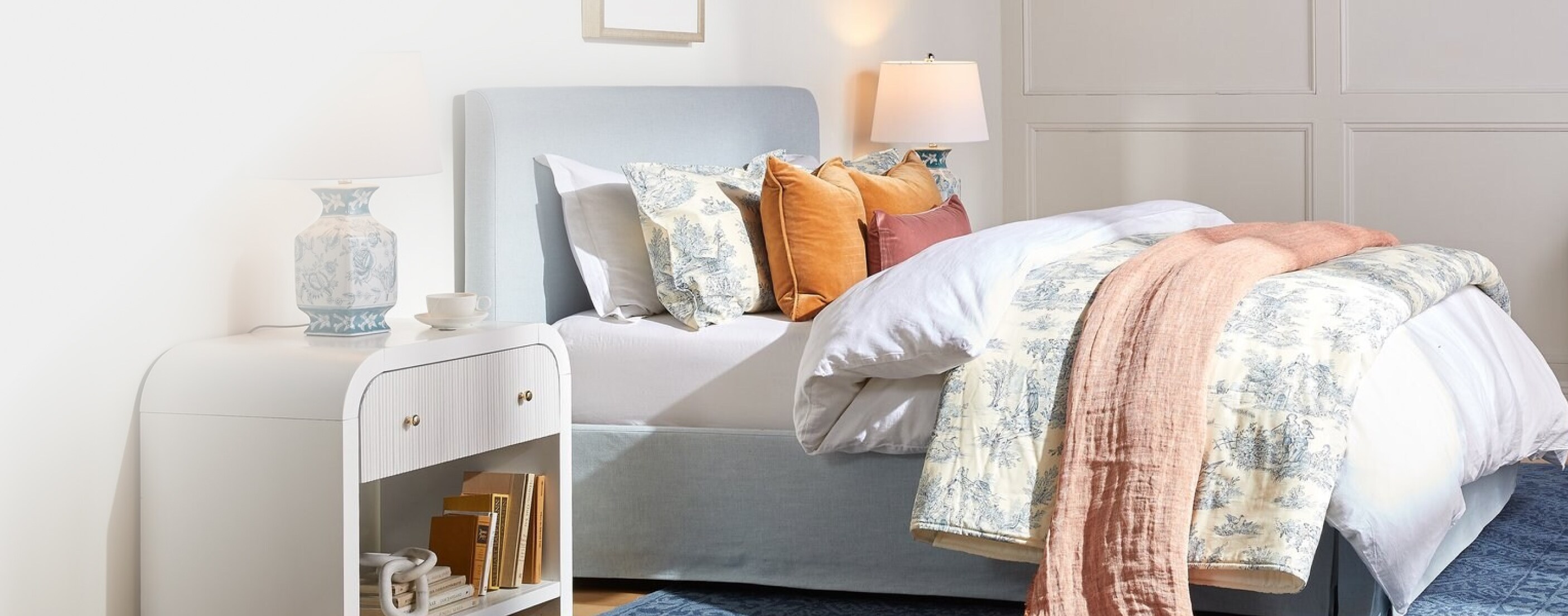 A light-blue upholstered bed is set with a plush white comforter, a patterned blanket, an orange coverlet and various pillows.
