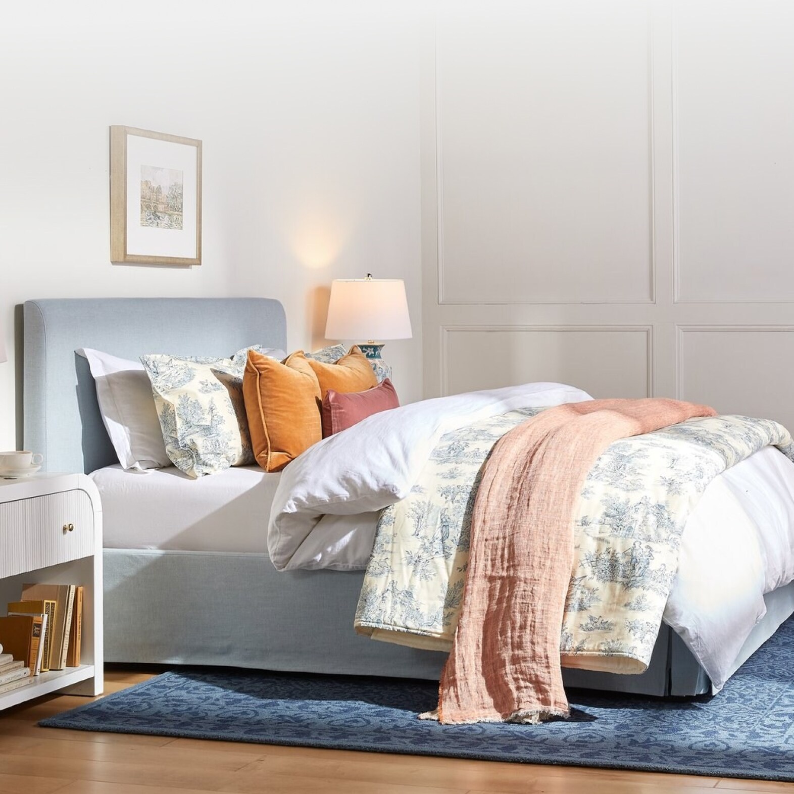 A light-blue upholstered bed is set with a plush white comforter, a patterned blanket, an orange coverlet and various pillows.