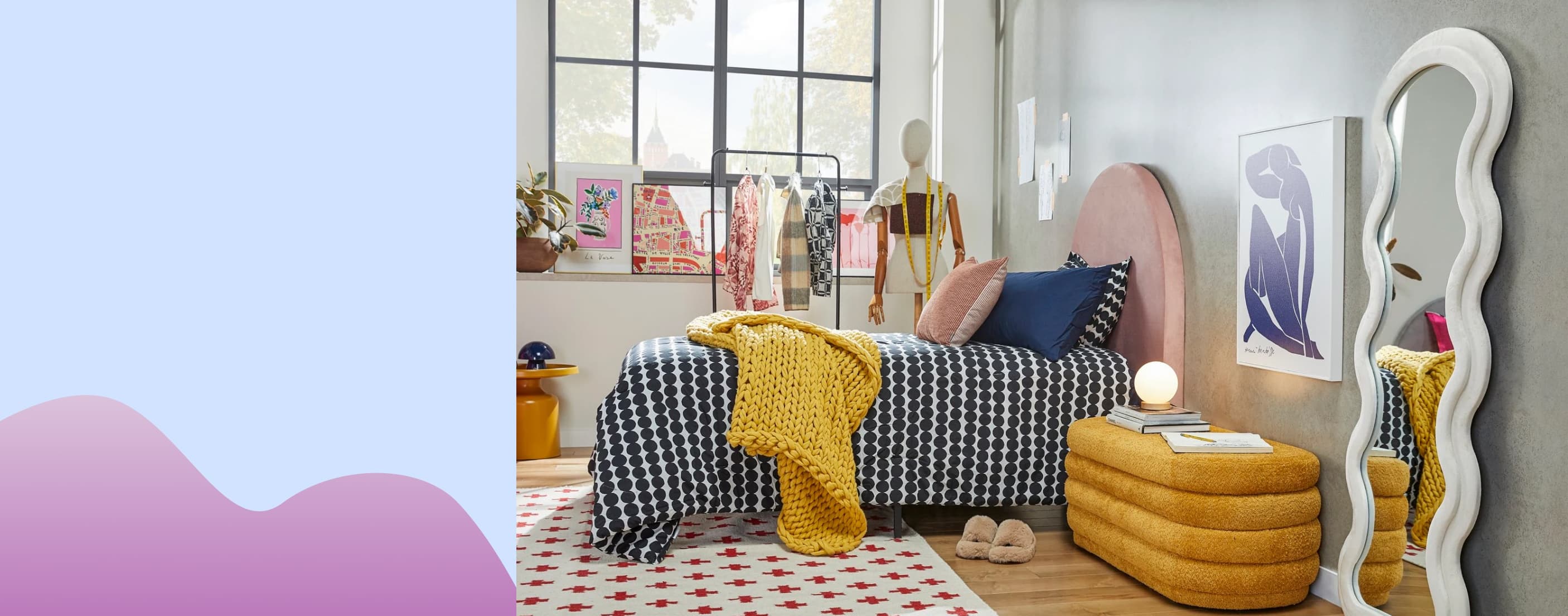 A trendy bedroom is shown with a unique curvy mirror, vevlet bench, artwork hung on the walls, clothes in the background, and more.
