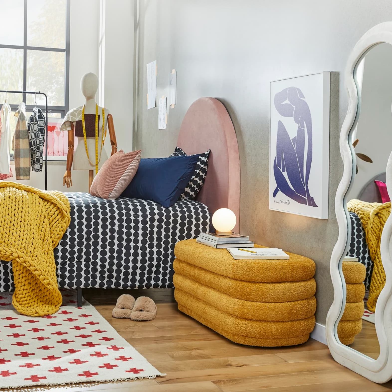 A trendy bedroom is shown with a unique curvy mirror, vevlet bench, artwork hung on the walls, clothes in the background, and more.