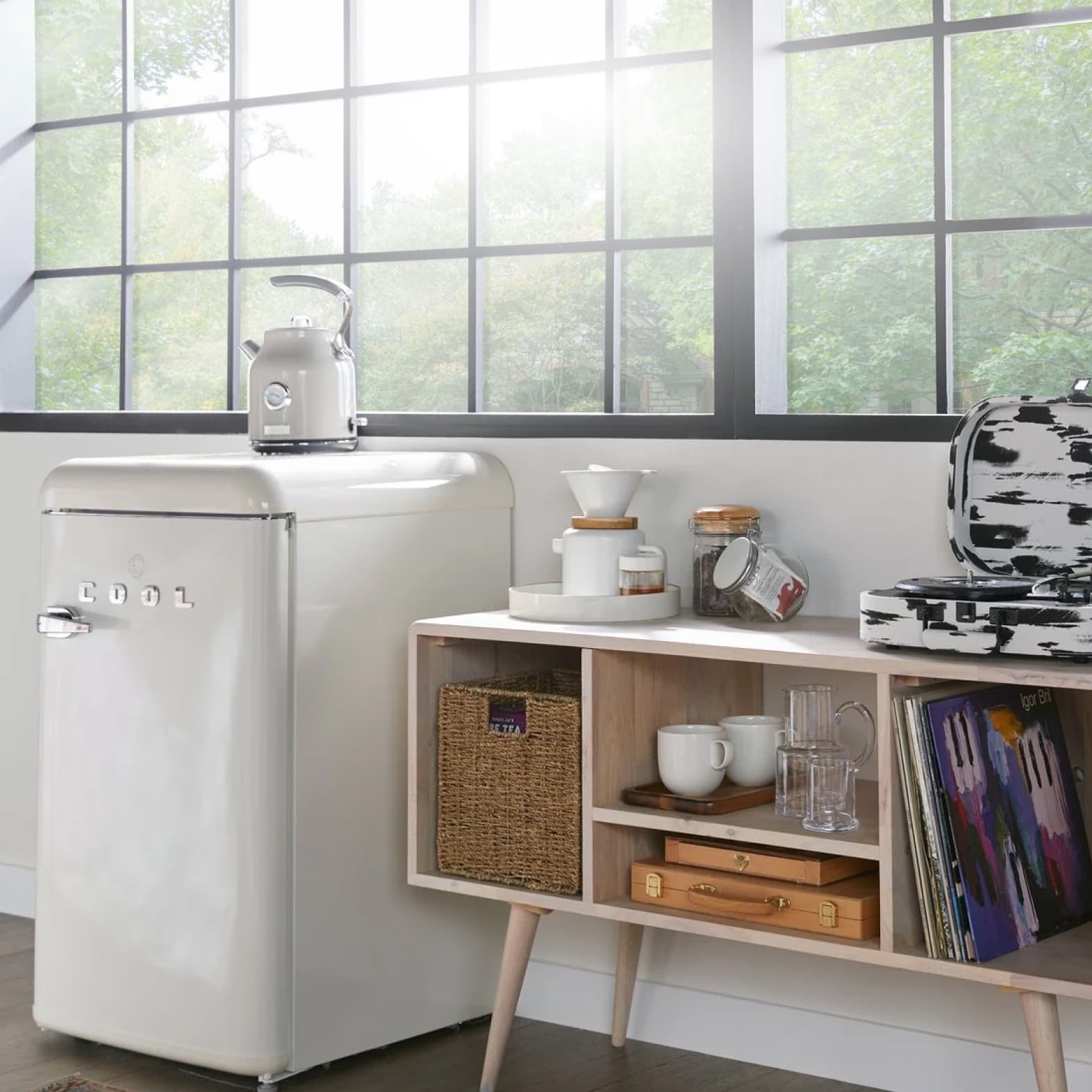 A white mini-fridge is shown with a coffee pot on top. Next to the fridge is a mid-century modern wooden console table with separated cabinets filled with books, dishes, and other supplies.