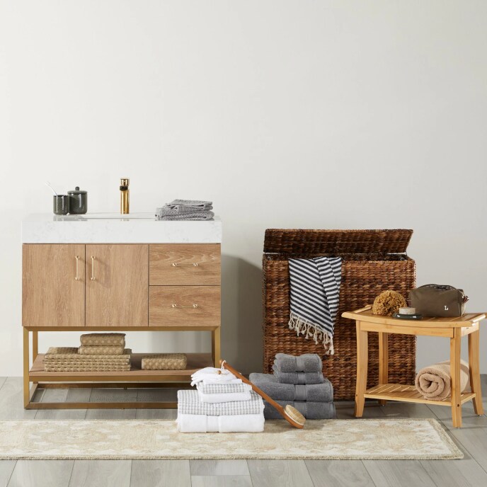 A light oak vanity with a white top and a brass faucet next to a woven wicker hamper, a wooden shower stool, and two stacks of towels.