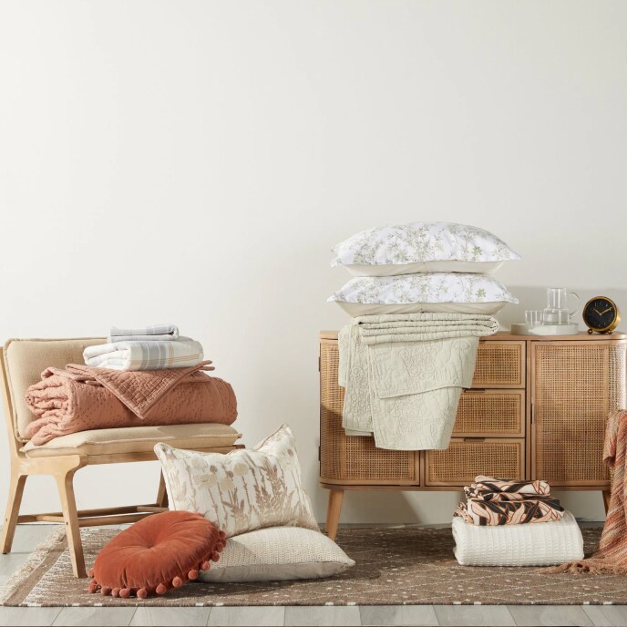 A mid-century modern slipper chair and a cabinet with a woven front, topped with and surrounded by pillows, sheets, and blankets. 