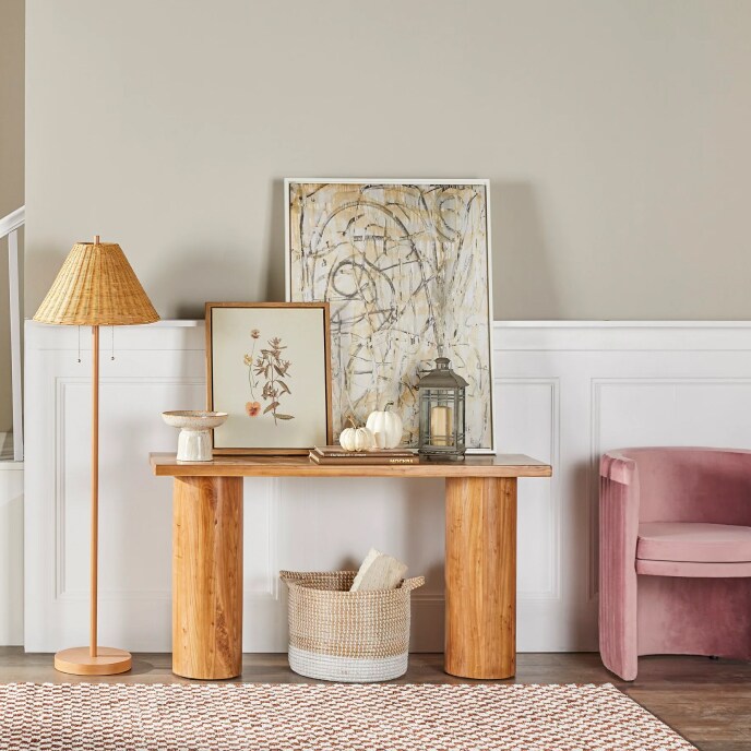 A wooden table with pillar legs, topped with artwork and set next to a velvet pink chair and a lamp with a wicker shade. 