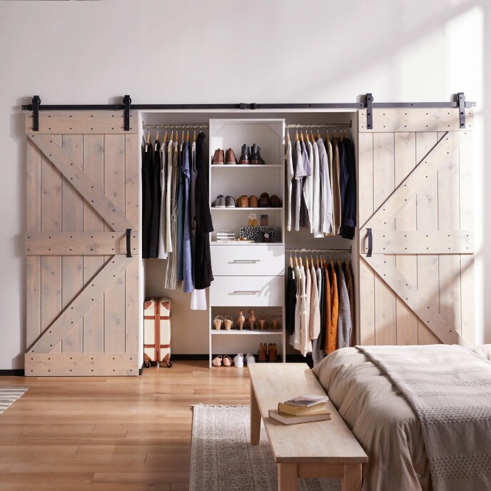 A closet with barndoor style doors that are opened to reveal neatly organized shoes and clothing in a bedroom with a wooden bench and a bed.