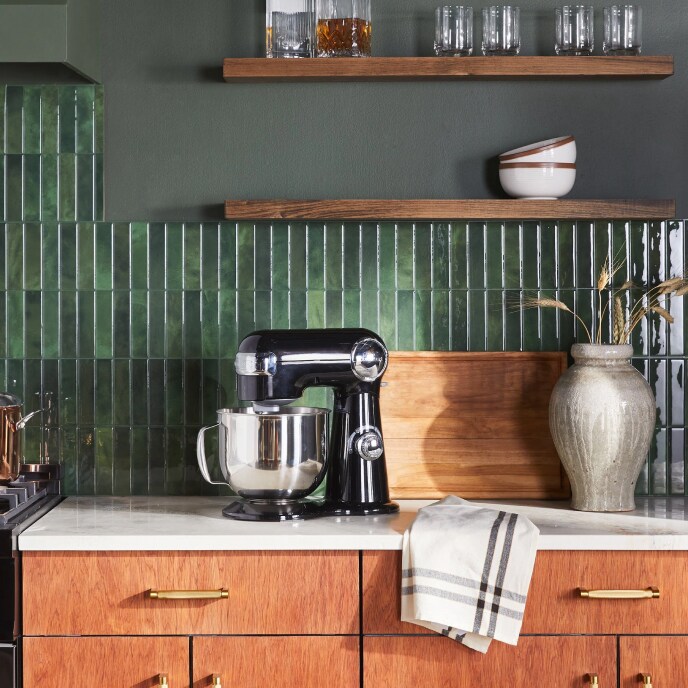 A black stand mixer and a ceramic vase on a white marble counter in a kitchen with wooden cabinets and a green tile backsplash. 