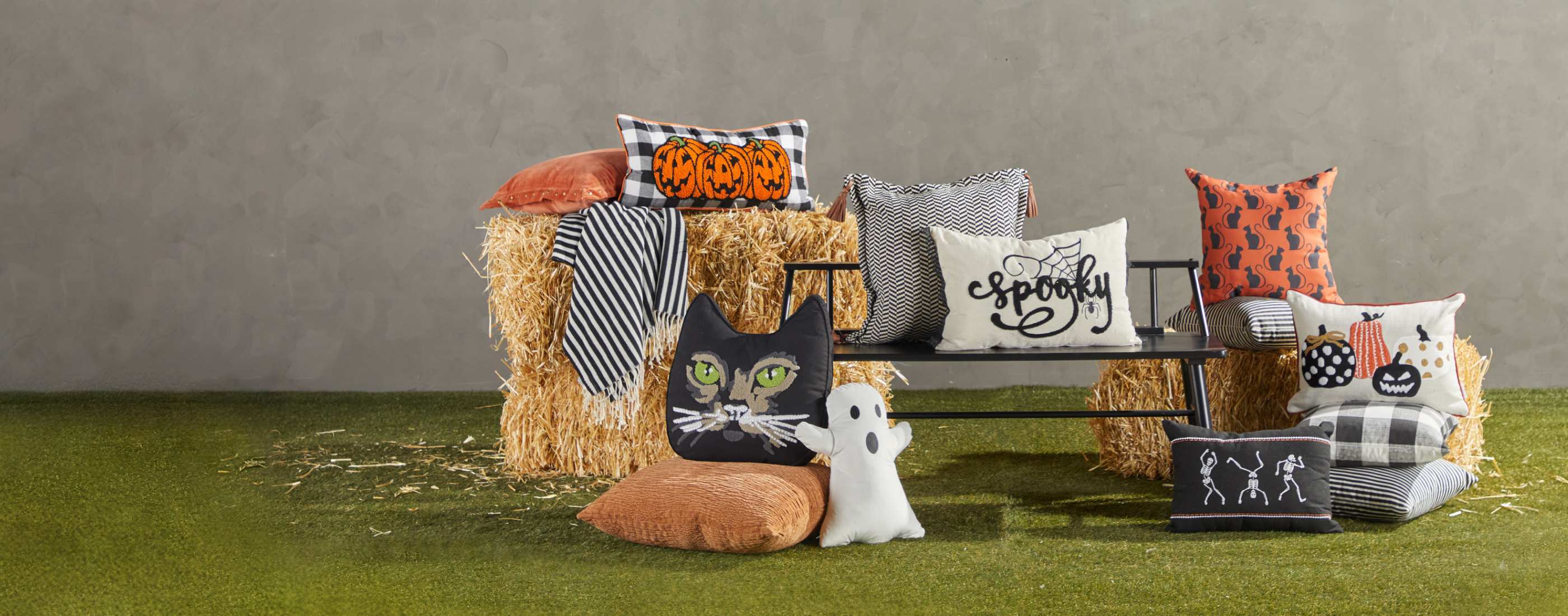 A halloween-themed bench with several spooky throw pillows including one shaped like a ghost, all in front of a bale of hay.
