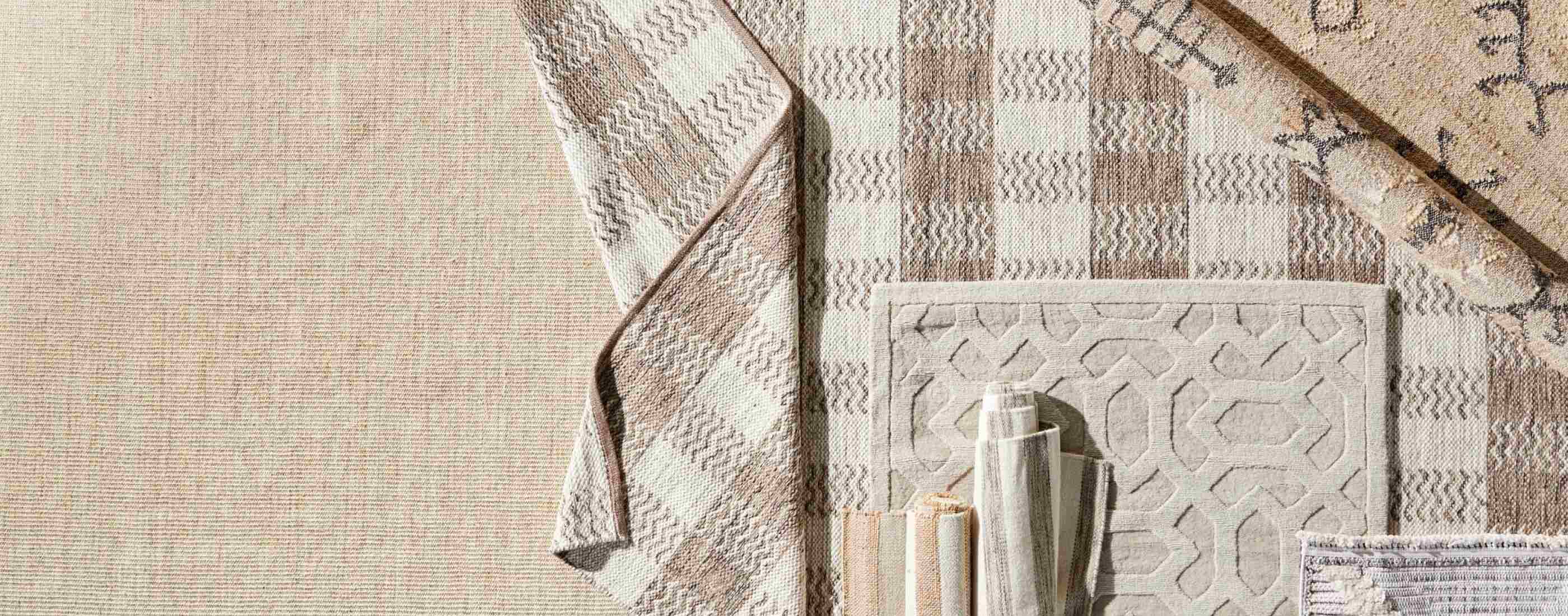 Assortment of rugs in beige and brown hues, some solid and some with a checkered pattern, layered and rolled out over each other.