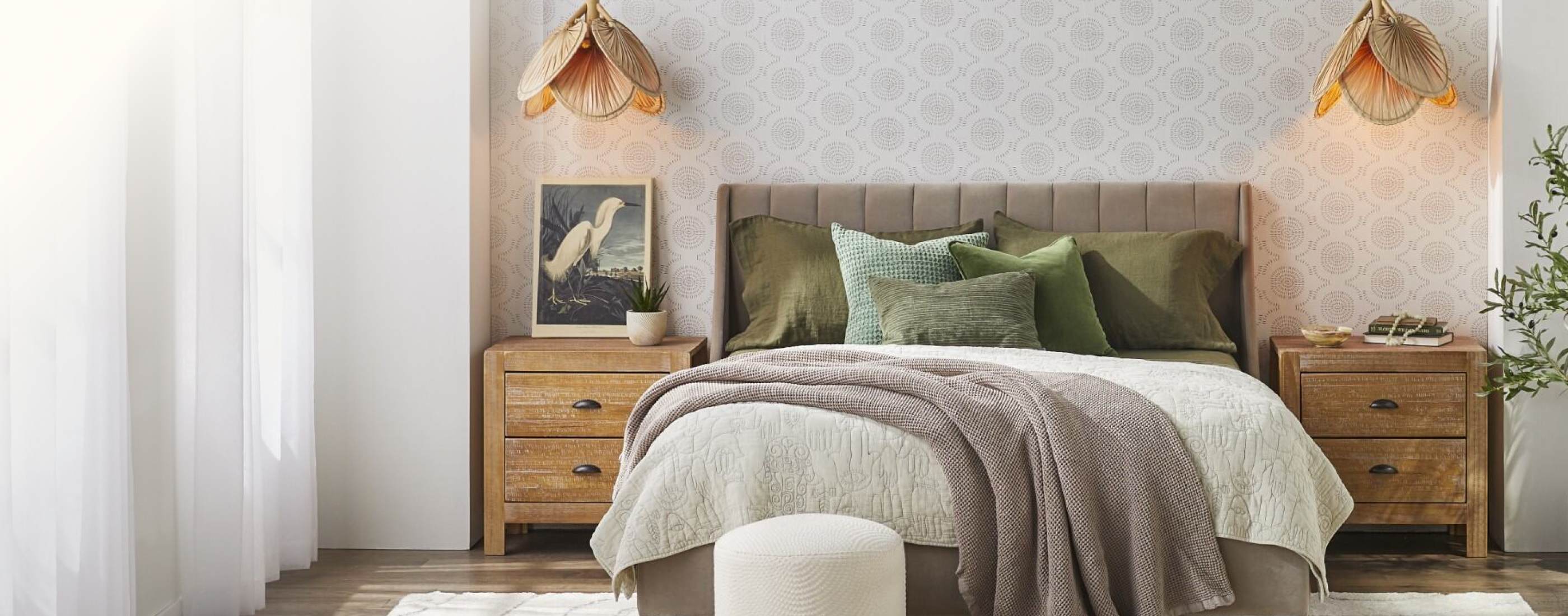 Coastal-style bedroom with natural wood nightstands, mattress layered with cozy blankets and pillows, and a tufted taupe velvet headboard.