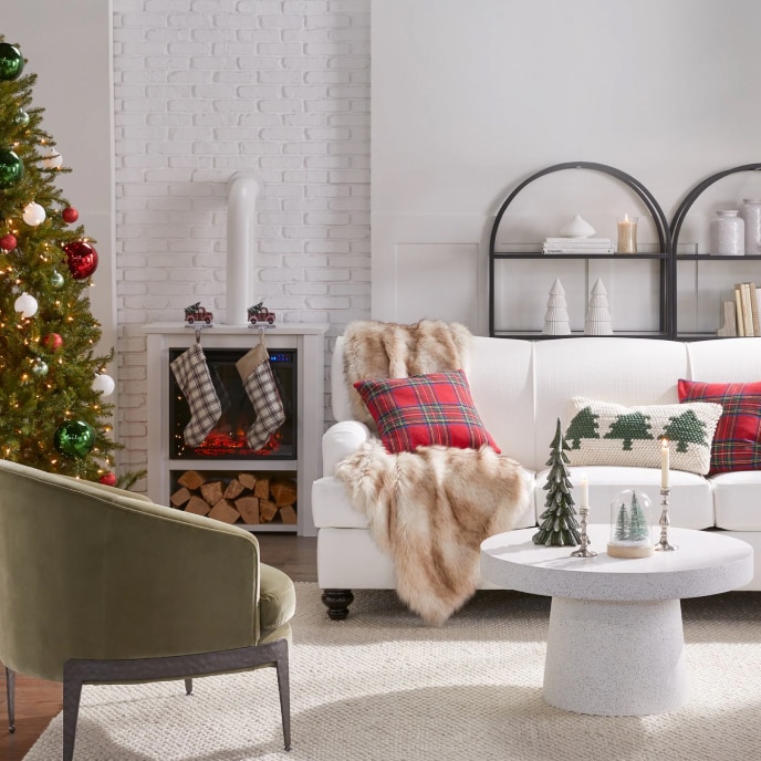 A cozy living room with a white sofa set with winter-themed throw pillows and a faux fur throw, next to a fireplace with stockings, a coffee table with Christmas décor, and a Christmas tree.