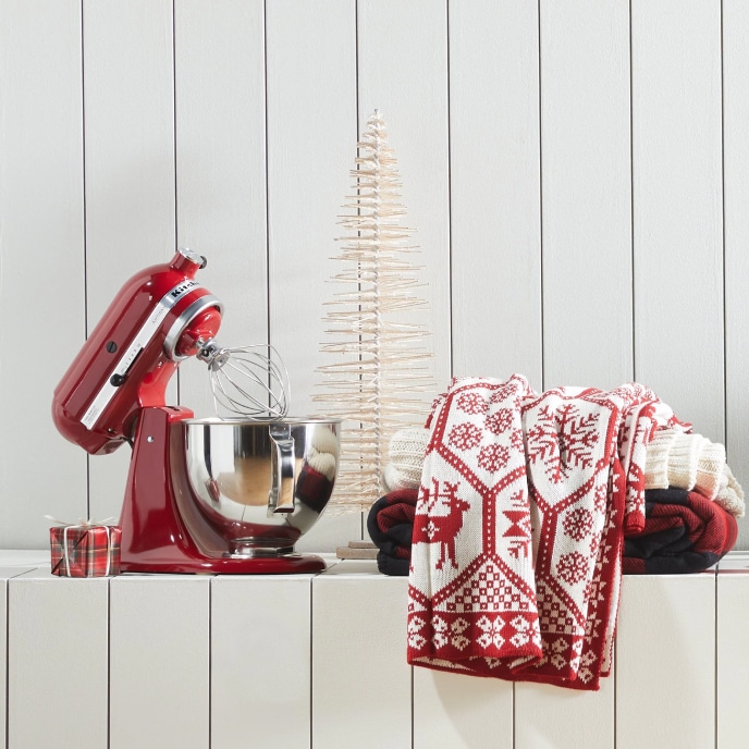 A display of giftable items, including a red KitchenAid mixer, a wrapped present, and a stack of red and white throw blankets next to a tabletop Christmas tree.