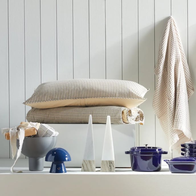 A display of giftable items, including a cozy pillow and blanket, a waffle-weave towel, decorative ceramics, pyramid-shaped marble sculptures, and a set of blue enamel cookware.