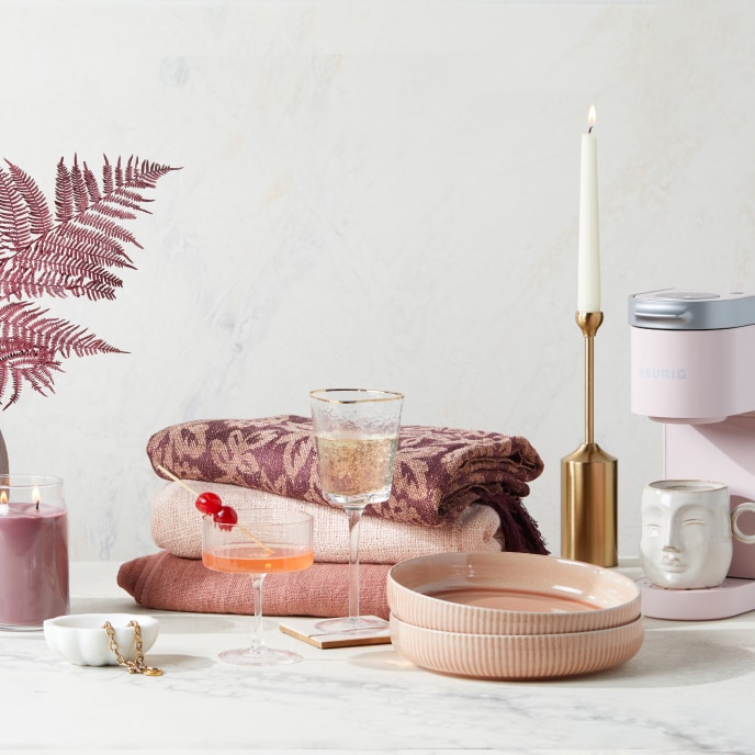 A set of giftable items including a pink candle, a pink fern, a stack of pink blankets, pink dishes, a pink coffee maker, a white ceramic jewelry dish, and cocktail and champagne glasses.