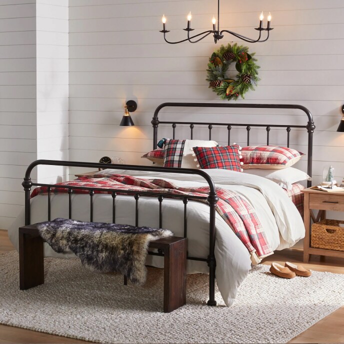 A cozy, black cast iron bed with red plaid and white bedding beneath a black candelabra chandelier and a wall with a pine wreath. 