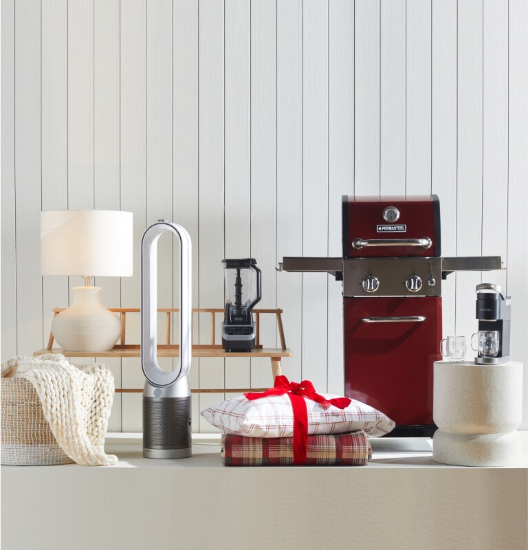 A variety of giftable products are displayed against a white backdrop, including a woven throw blanket, table lamp, blender, outdoor grill, bedding, and tabletop mixer.