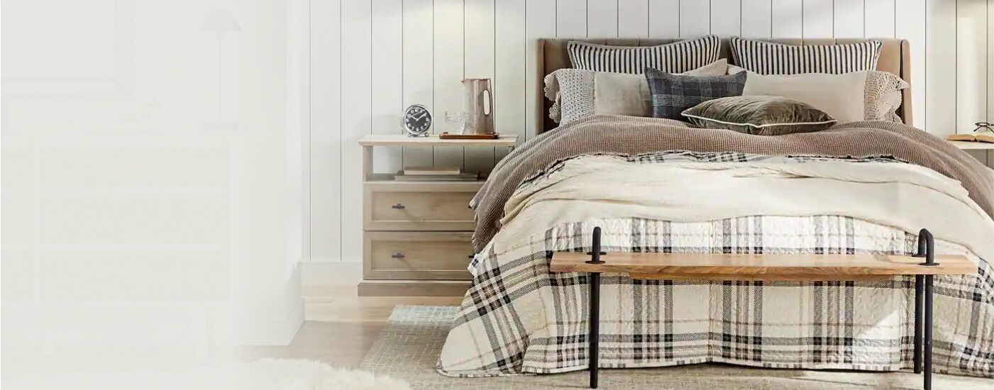 An inviting bed, nightstand, and bench are displayed. The bed is layered with various cozy pillows and blankets, with a brown-and-white checkered blanket on top.