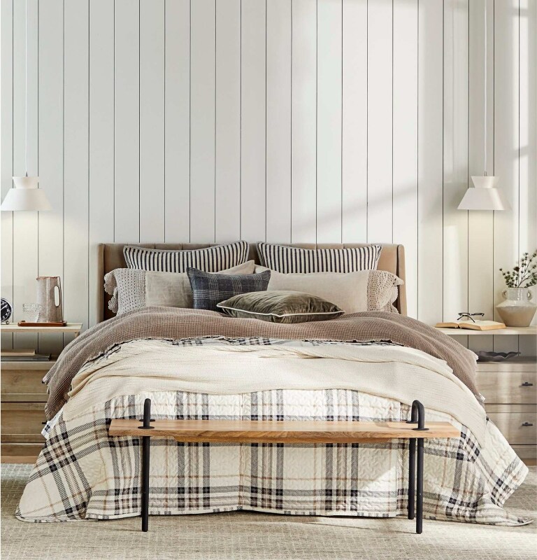 An inviting bed, nightstand, and bench are displayed. The bed is layered with various cozy pillows and blankets, with a brown-and-white checkered blanket on top.