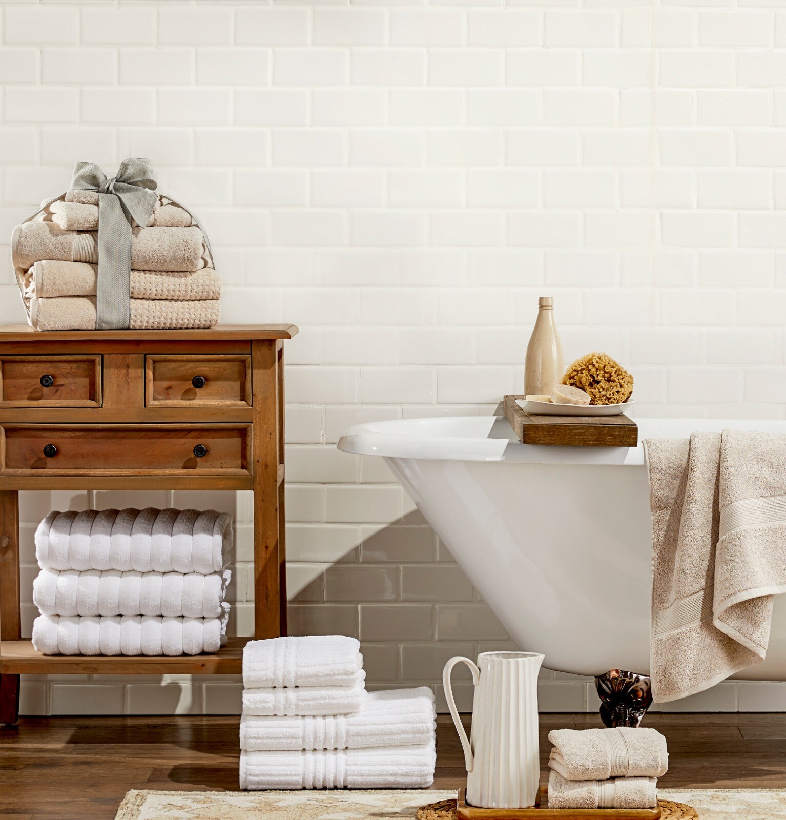 A bathroom with stacks of beige and white towels on a wooden side table next to a stack of towels and a pitcher on the floor, and a clawfoot tub with a wooden bathtub caddie.