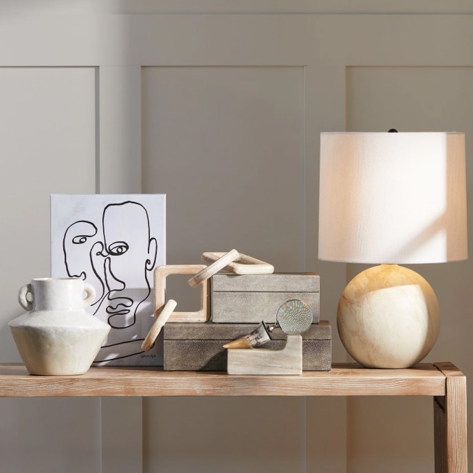 A console table set with a vase, an abstract line drawing of a man’s face, wooden chain links laid over decorative boxes, a horn-handle magnifying glass, and a lamp with a spherical base.