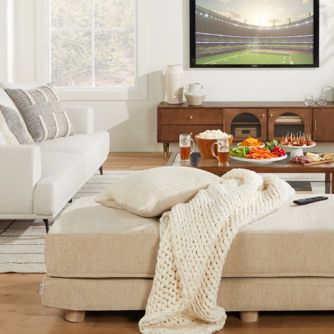 A rectangular, beige ottoman covered with a pillow and a knit blanket in a living room with a white sofa, a wooden coffee table covered with snacks, and a TV showing a football field.