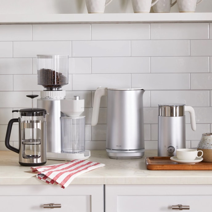 A white kitchen counter displaying a variety of coffee and tea machines and accessories, including a French press, a coffee grinder, a large and a small electric kettle, and a wooden serving tray with a teacup.