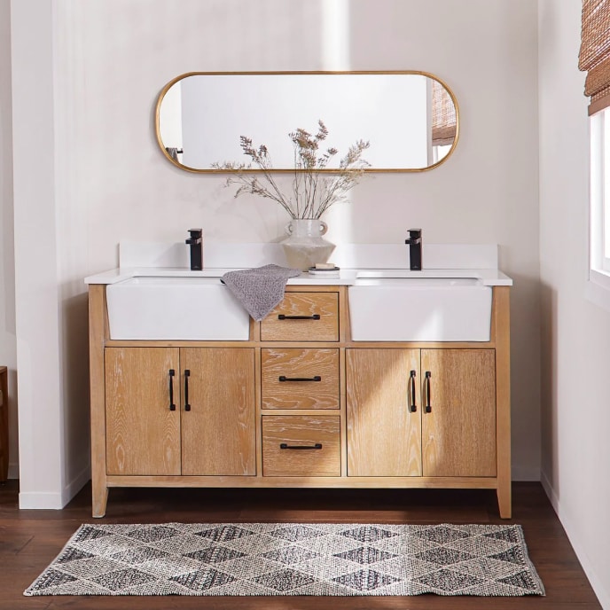 A double sink, natural wood bathroom vanity with drop-in apron sinks and oil-rubbed bronze hardware beneath a gold-framed oval mirror.