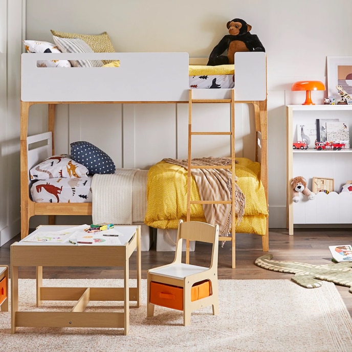 A kid’s bedroom with white and natural wood bunk beds with safety rails, a play-room table, a book shelf, and a variety of stuffed animals and kid’s toys.