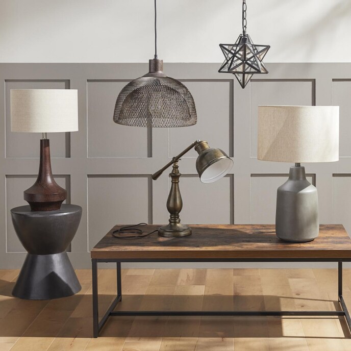 A display of modern lighting including a rust-colored lamp on a grey pedestal table, a metal shade chandelier, a star-shaped chandelier hanging from the ceiling, and a wood table with two lamps.