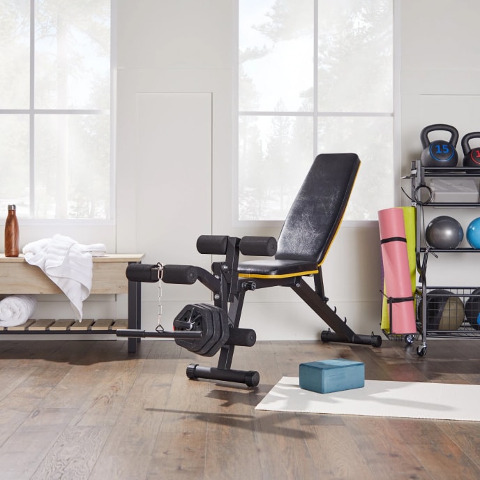 A home gym with a leg weight lifting machine, a yoga mat and block, a weight shelf, and a wooden bench with towels.