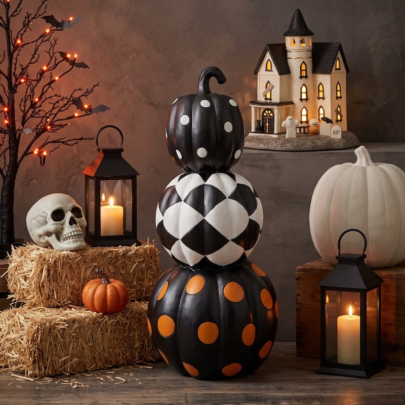 Halloween decor display featuring three black, white, and orange stacked pumpkins, a lit haunted house, glowing lanterns, a skull, hay bales, and pumpkins.