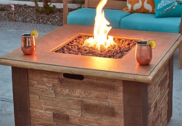 Lit propane fire pit with stone sides beside a patio bench with turquoise cushions